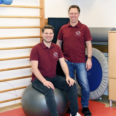 Jens-Peter Markhoff und sein Sohn Lasse stehen bzw. sitzen vor Trainingsgeräten, wie der Sprossenwand und einem Trampolin und lächeln in die Kamera.