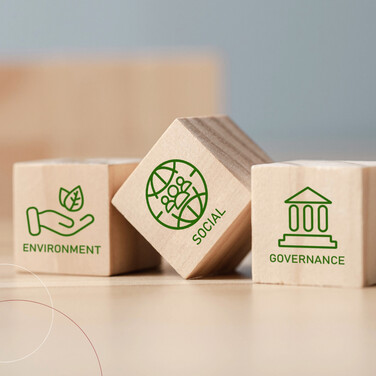 Drei Holzwürfel liegen auf einem Tisch. Jeder Würfel hat ein Wort und ein Bild auf der Frontseite. Würfel 1: Environment mit einer Hand und Blättern darüber; Würfel 2: eine Weltkugel mit Menscehn, darunter das Wort Social; Würfel 3: ein Säulengebäude grafisch dargestellt, darunter das Wort Governance.